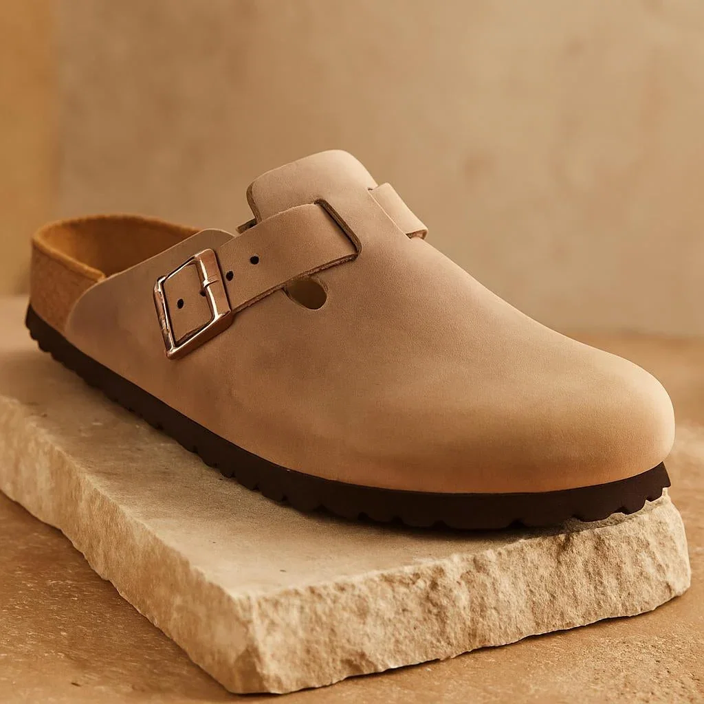 Beige leather clog with buckle strap on dark sole, displayed on stone platform against tan background
