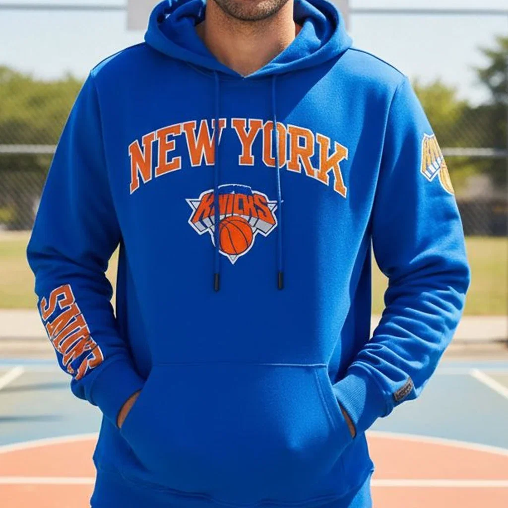 Man wearing blue New York Knicks hoodie with orange text and basketball logo on outdoor court