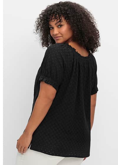Ein Model trägt eine schwarze Kurzarmbluse mit leicht transparente Lochstickereien, Carmenausschnitt, Volant und Bindeban