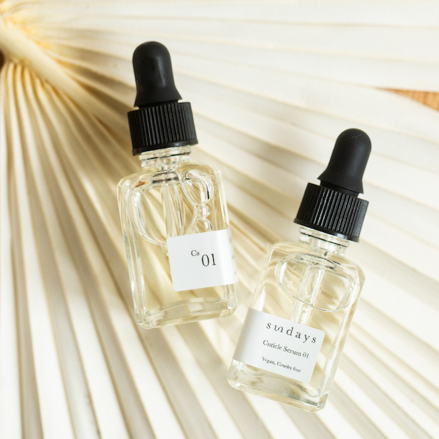 Two bottles of cuticle serum with droppers on a neutral palm leaf background.