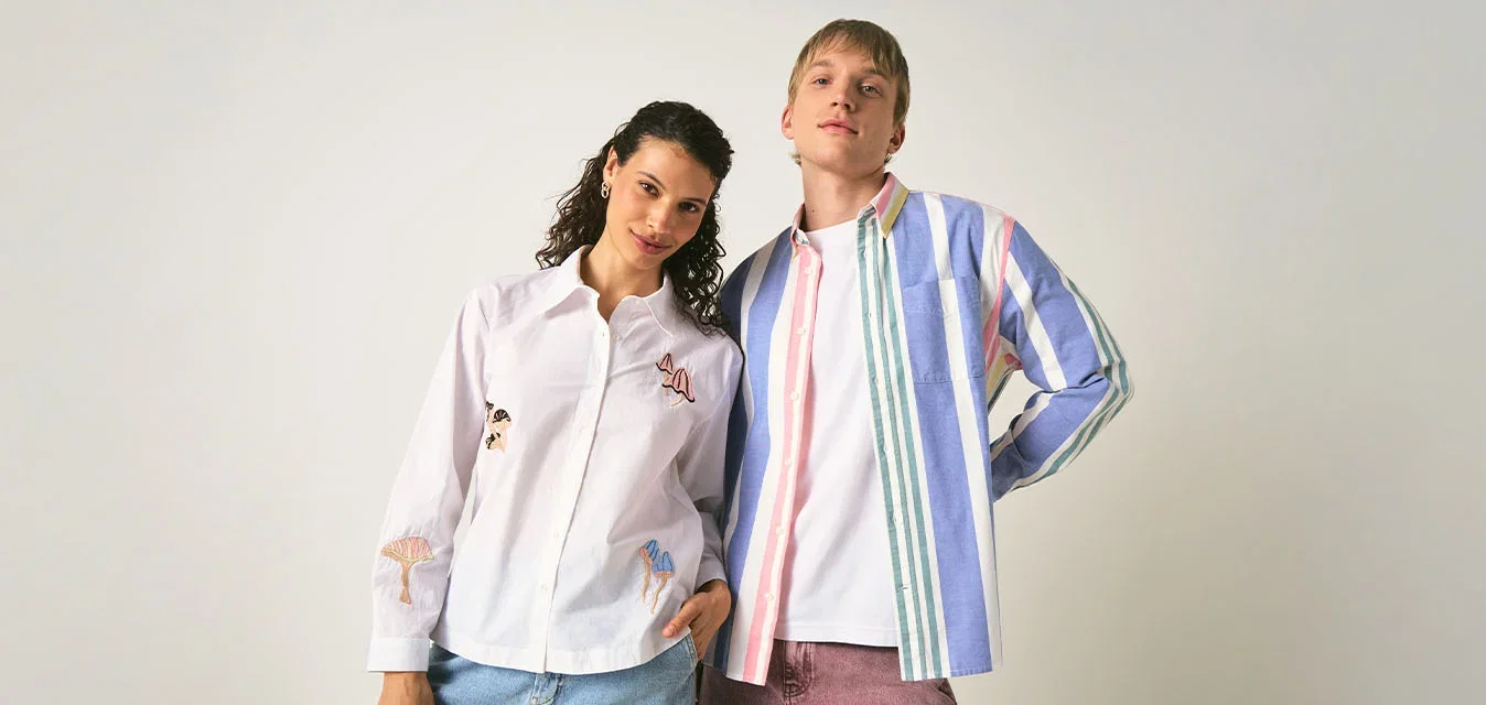 Dos jóvenes adultos vestidos de forma informal están uno al lado del otro contra un fondo claro y liso, uno lleva una camisa a rayas y el otro una camisa blanca con diseños.