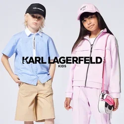 Dos niños están de pie; uno lleva una camisa azul de rayas con pantalones cortos beige y una gorra negra, el otro lleva un chándal rosa y un sombrero con un pequeño bolso.