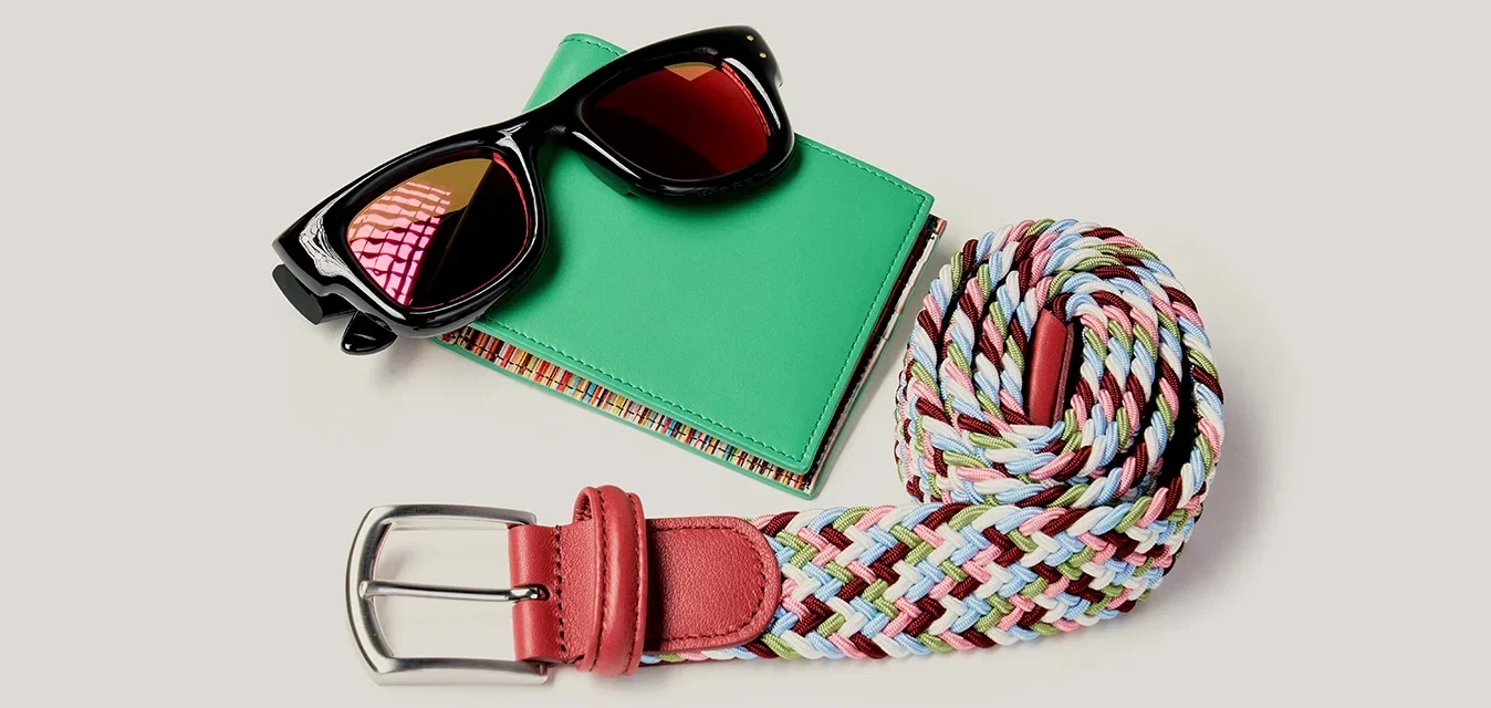 Gafas de sol negras sobre una cartera verde junto a un cinturón tejido multicolor enrollado con una hebilla de cuero roja sobre un fondo claro.