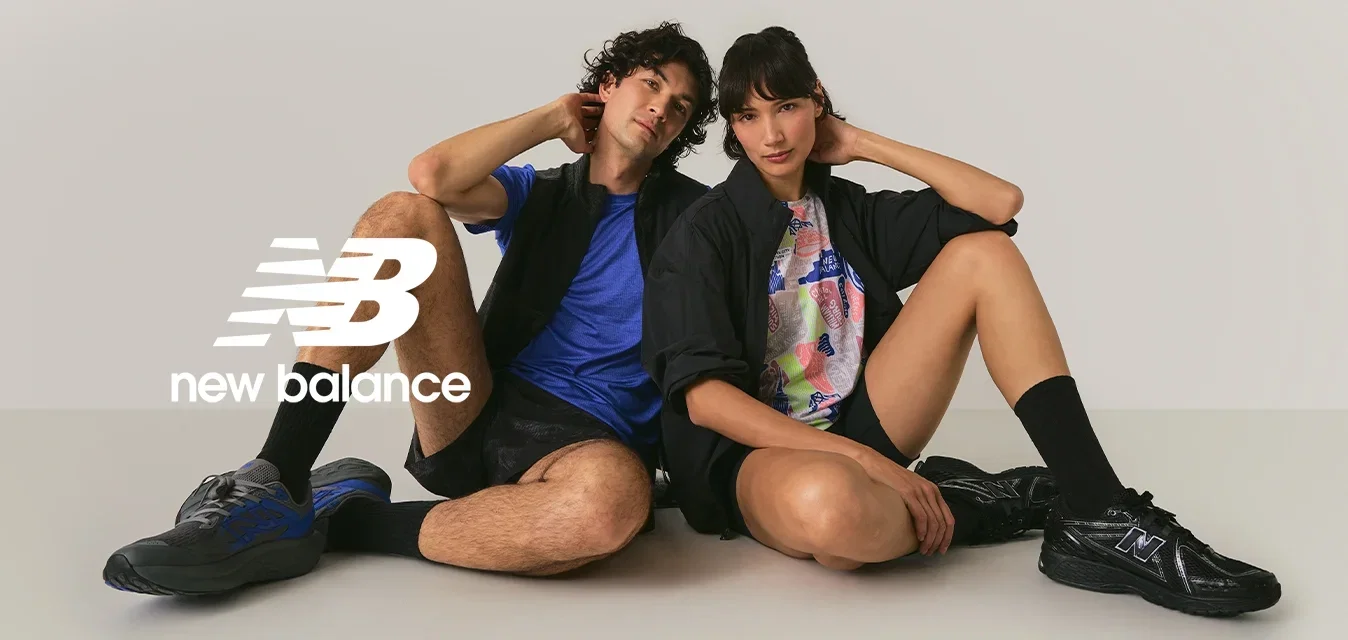 Dos jóvenes sentados en el suelo con las piernas dobladas, vistiendo ropa deportiva casual y zapatillas, frente a un fondo claro y sencillo.