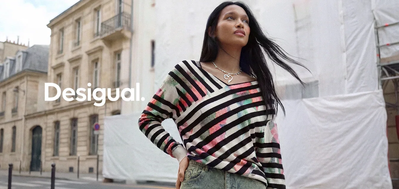 Mujer con cabello largo y oscuro que lleva un jersey de rayas multicolor y una falda vaquera, de pie en una calle de la ciudad con edificios históricos.