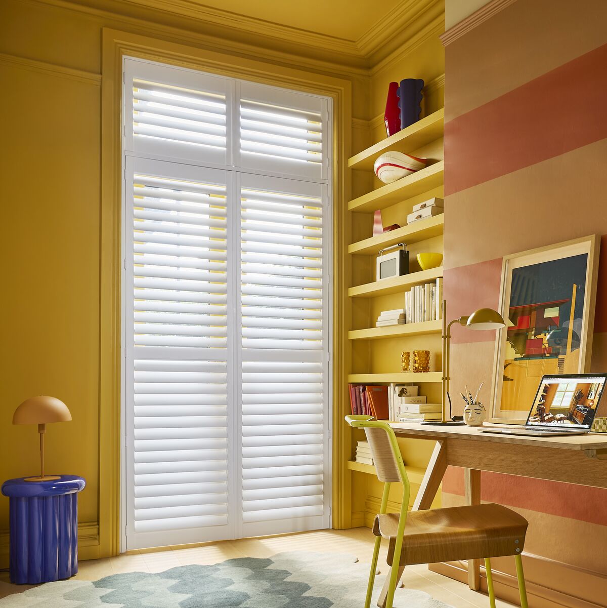 White shutters cover a tall window beside a desk, bringing soft light to a colourful home office with striped walls and shelving.