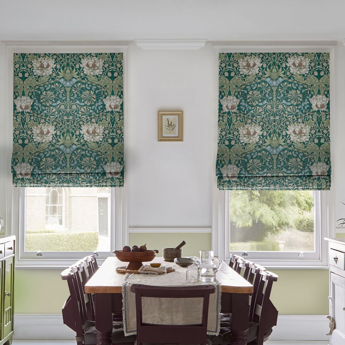 Pair of floral Roman blinds in teal and cream frame tall dining room windows, bringing pattern and softness to the light-filled space.