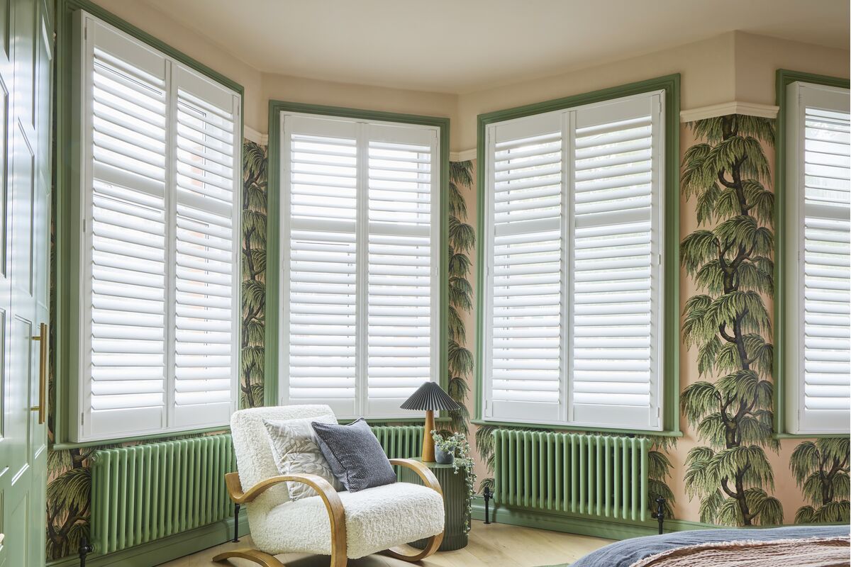 White shutters span three bay windows, filtering natural light into a bedroom with botanical wallpaper and soft green trim.