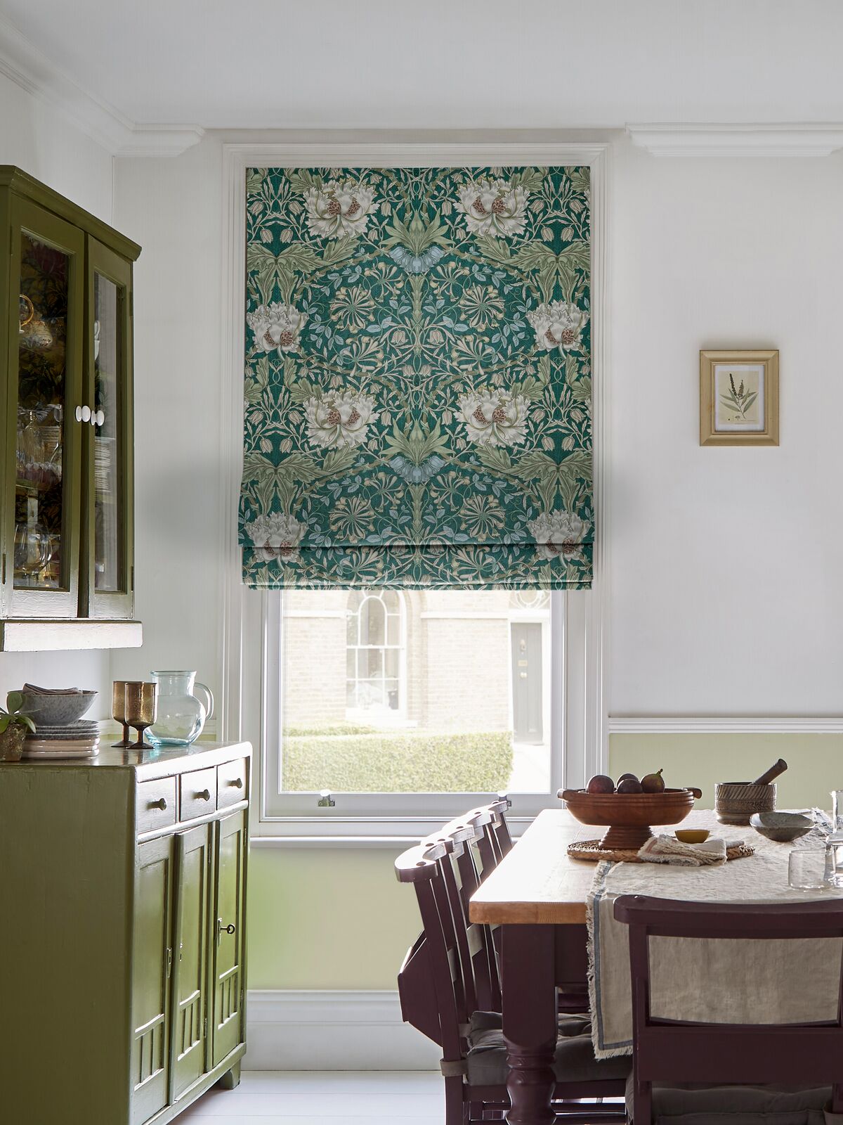 Pair of floral Roman blinds in teal and cream frame tall dining room windows, bringing pattern and softness to the light-filled space.