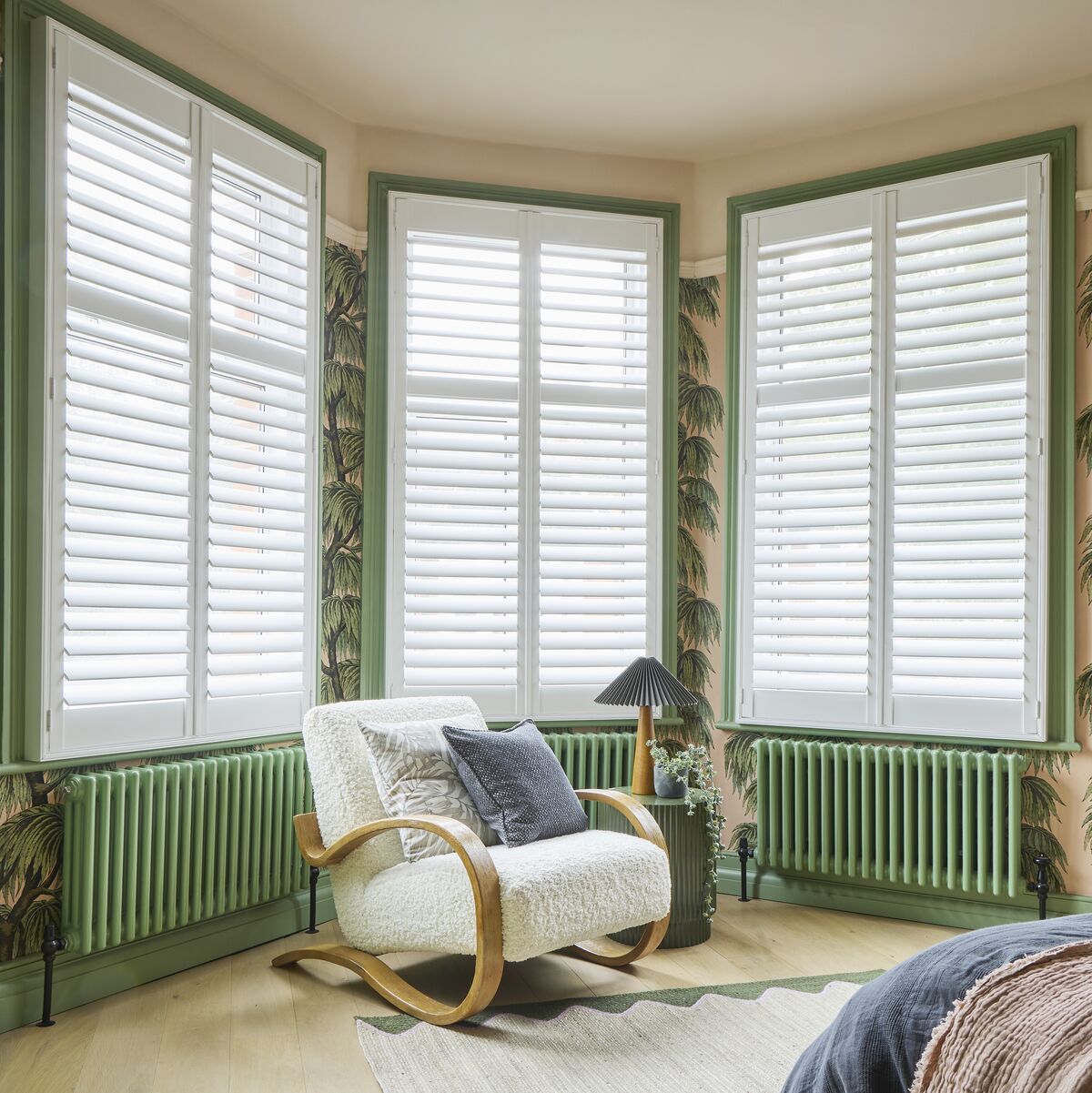 White shutters span three bay windows, filtering natural light into a bedroom with botanical wallpaper and soft green trim.