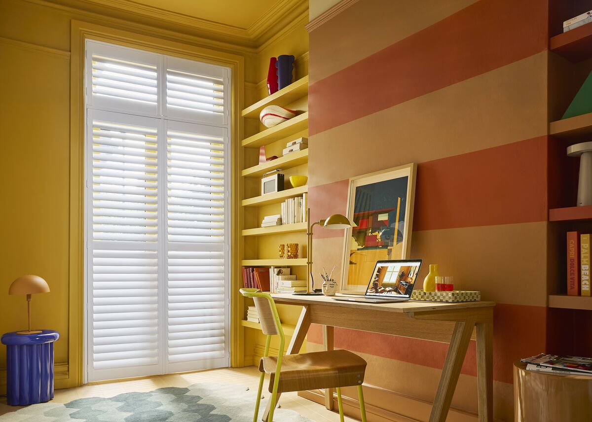 White shutters cover a tall window beside a desk, bringing soft light to a colourful home office with striped walls and shelving.