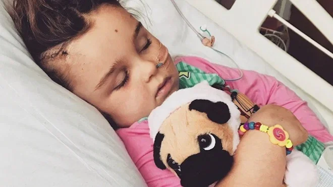 Lilly-May, a young girl, in a hospital bed, cuddling a soft toy dog while recovering from brain surgery.