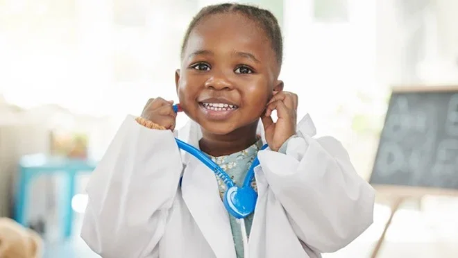 Smiling young child wearing a doctor's coat and holding a blue toy stethoscope to her ears.