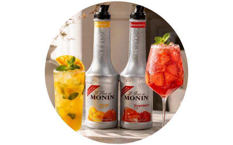Two Monin fruit syrup bottles (Mango, Strawberry) flanked by a mango mint iced drink and a strawberry cocktail garnished with fresh slices and mint