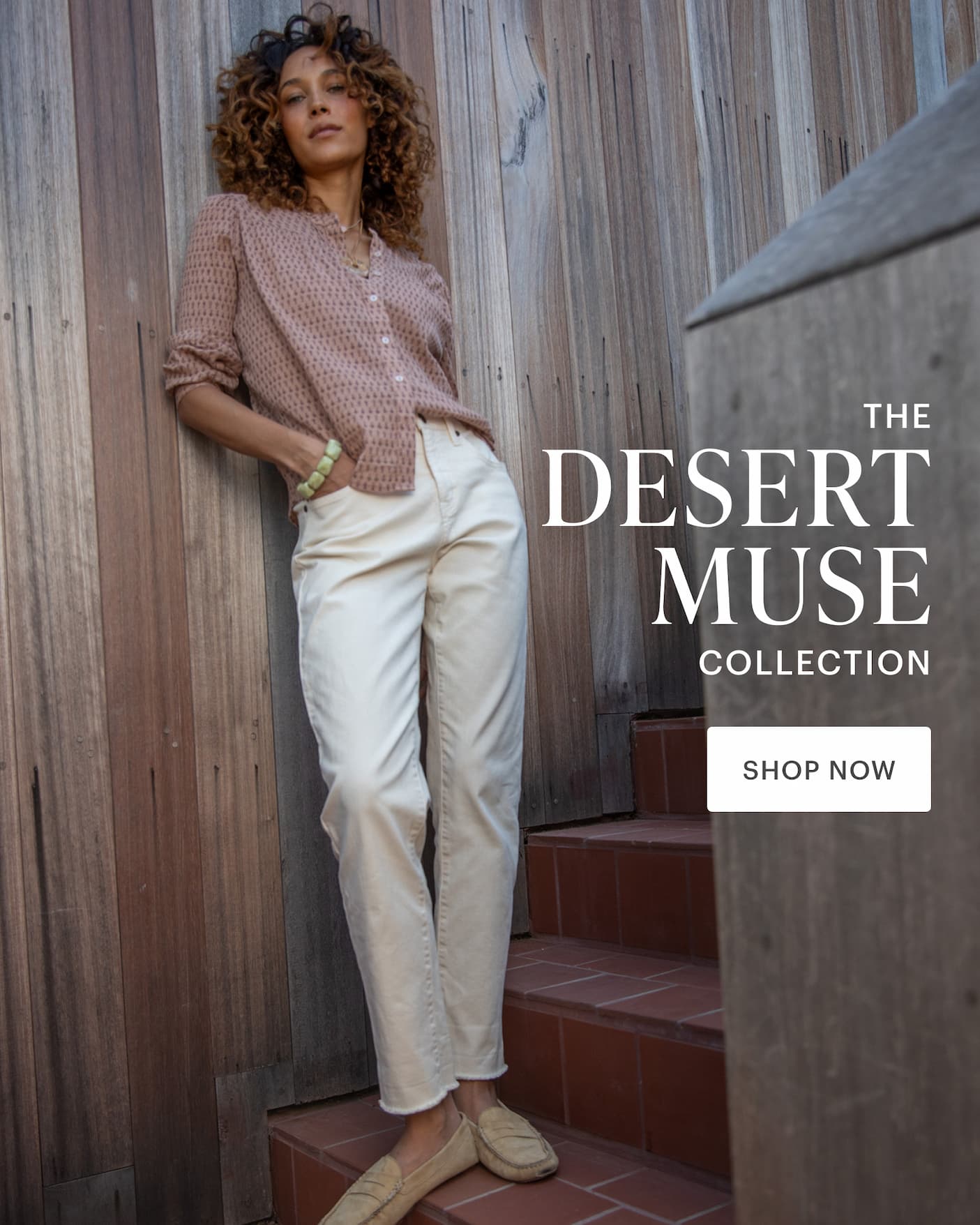Woman in elegant button up top with casual off-white pants, standing by wooden wall. 