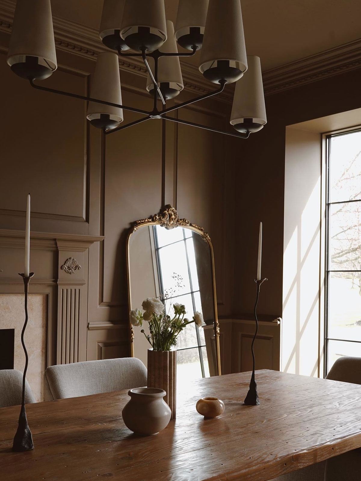 Elegant taupe dining space with wooden table, gold mirror, and sculptural chandelier.