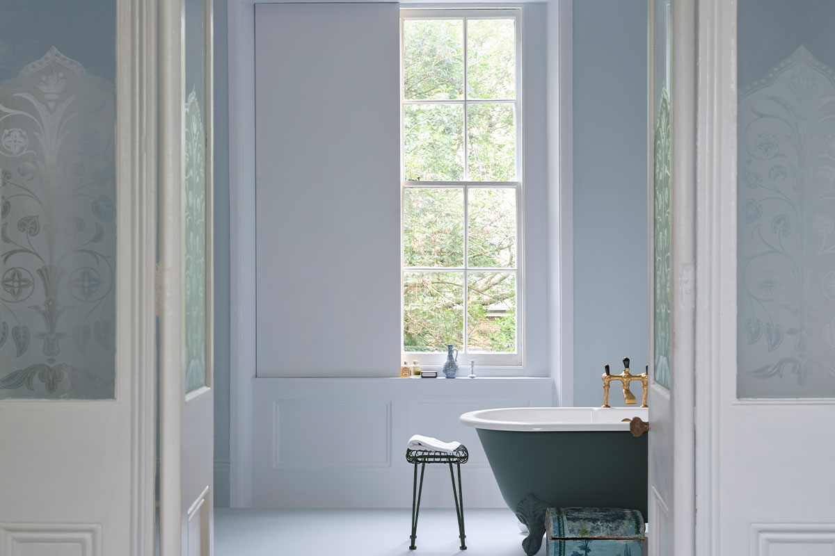 Calm blue bathroom with freestanding dark tub, gold taps and large window overlooking greenery.