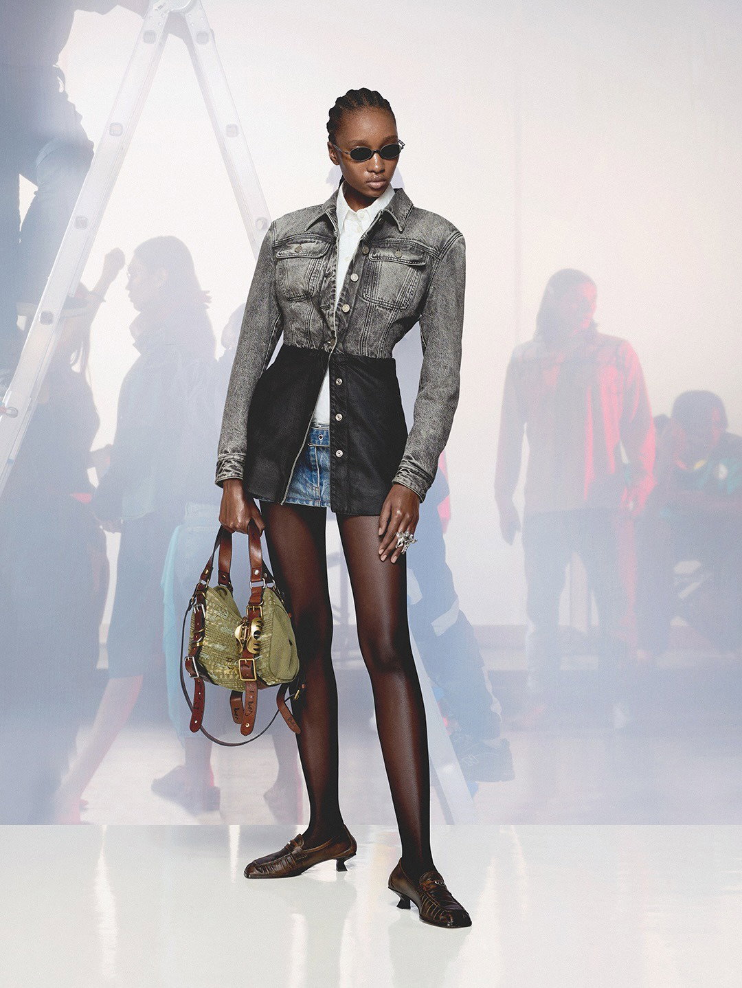 Studio fashion shot: model in two-tone denim jacket, mini skirt and black tights, sunglasses and green handbag