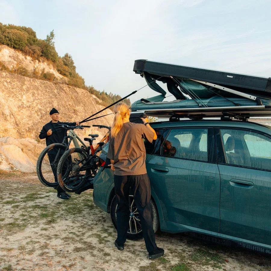 Two people preparing a vehicle-mounted roof tent at an outdoor campsite, with bicycles secured to the rear of the car and rocky hills in the background.