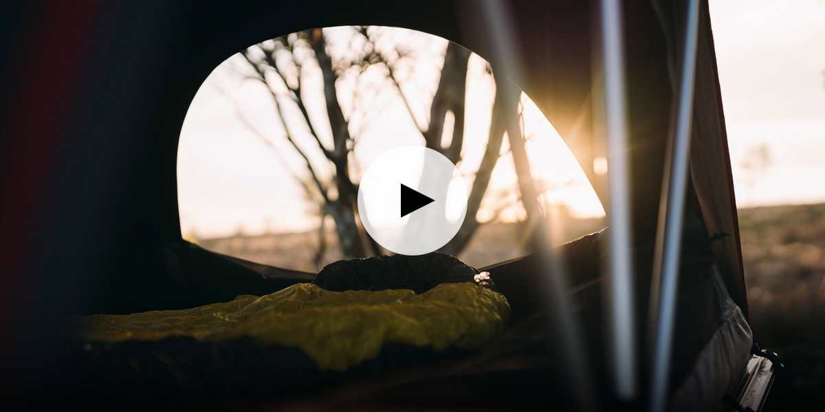 View from inside a roof tent showing a sleeping bag and a large window opening onto an outdoor landscape at sunrise. A play button icon is overlaid, indicating video content.