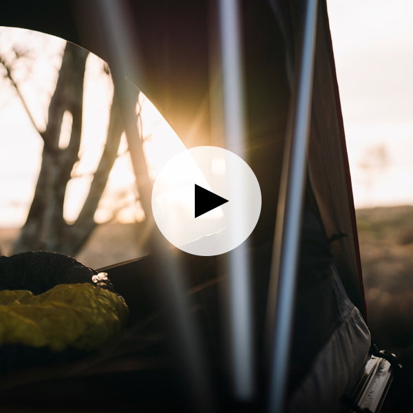 View from inside a roof tent showing a sleeping bag and a large window opening onto an outdoor landscape at sunrise. A play button icon is overlaid, indicating video content.