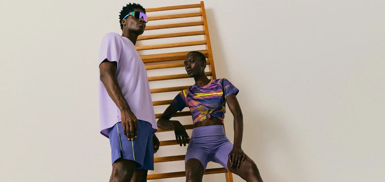 Zwei schwarze Models in sportlichen violetten Outfits posieren vor einer Holzleiters im hellen, minimalistischen Raum.