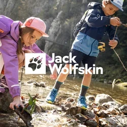 Zwei Kinder mit Rucksäcken spielen an einem felsigen Bach und legen dabei Stöcke und Blätter ins Wasser, im Hintergrund Wald und Felsen.