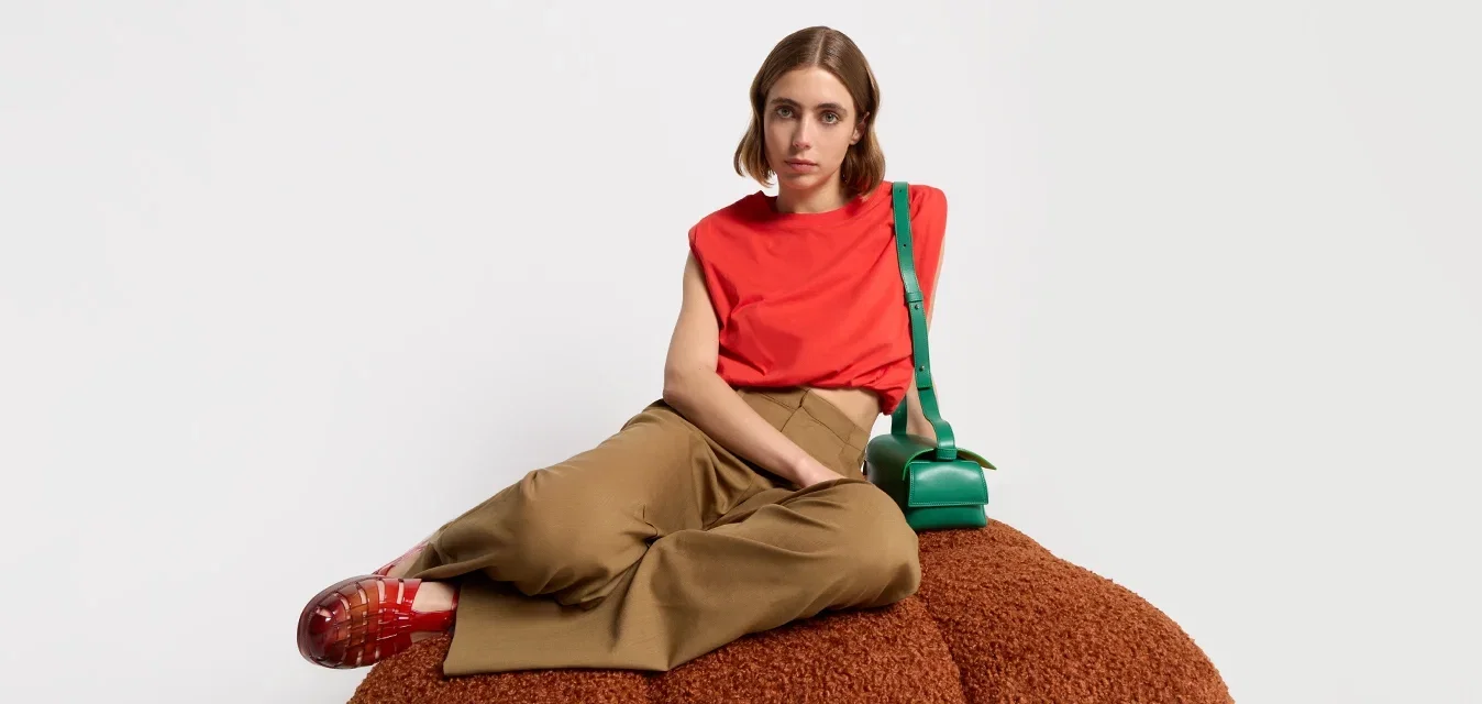 Pouf orange, texturé ; une personne portant une chemise rouge et un pantalon marron est assise à côté d'un sac vert ; arrière-plan minimaliste et lumineux.
