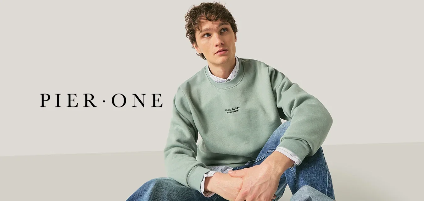 Jeune personne aux cheveux bouclés assise en tailleur, portant un sweat-shirt vert clair et un jean bleu retroussé, sur un fond uni.