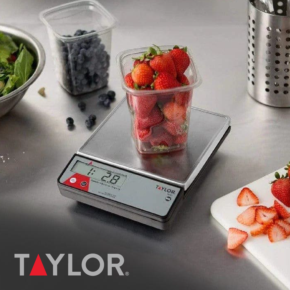 Taylor digital food scale weighing a container of fresh strawberries on a stainless steel countertop, with blueberries and additional produce containers nearby.