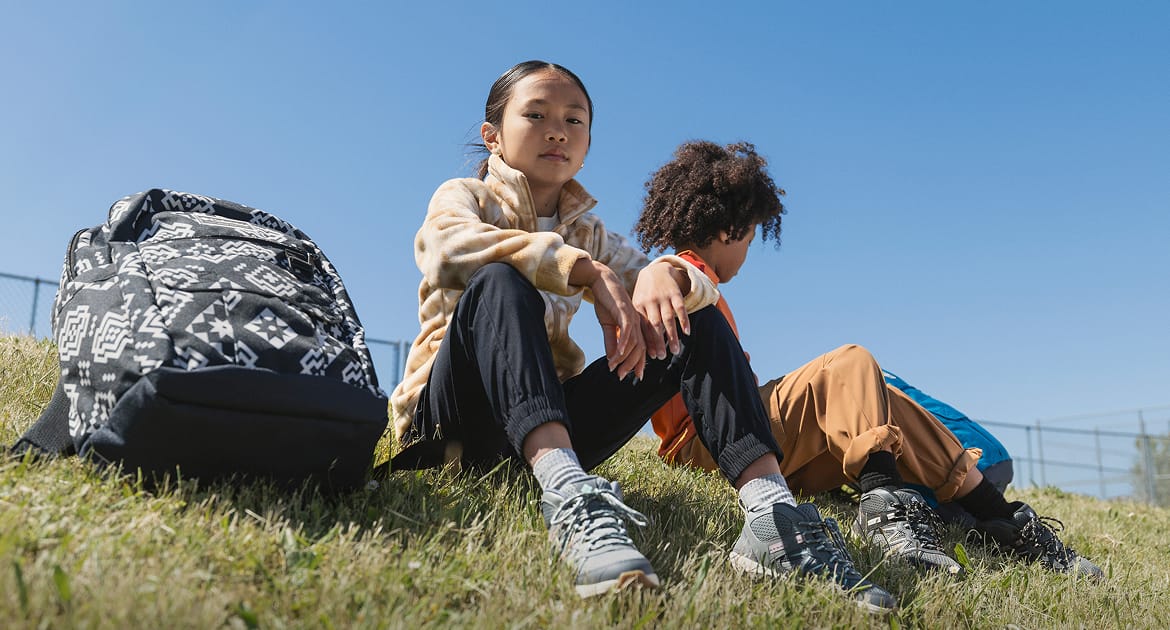 Two kids sitting on grass with backpacks.