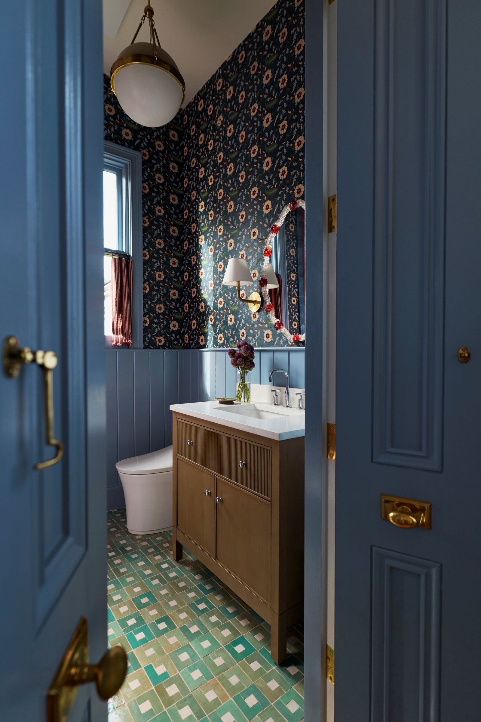 Dabito’s eclectic guest bathroom with blue paneled walls, floral wallpaper, patterned tile floor, and KOHLER vanity, faucet, and smart toilet viewed through an open door.