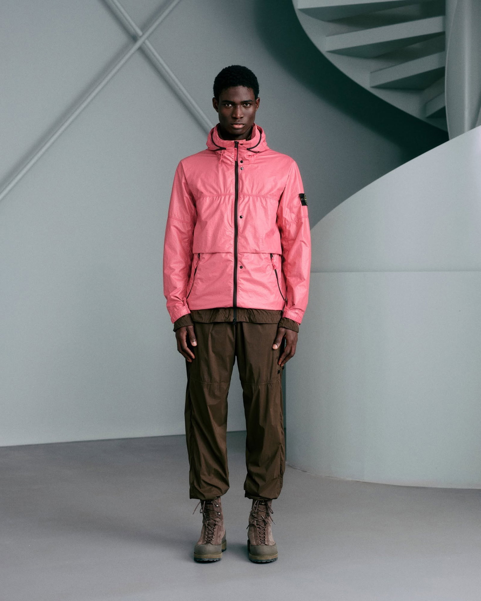 Male model wearing a pink hooded jacket, brown cargo trousers and hiking boots, standing in a minimalist studio with a curved staircase