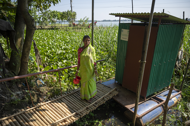 a woman stood on a bamboo gangway over wetlands, leading to an outdoor toilet.