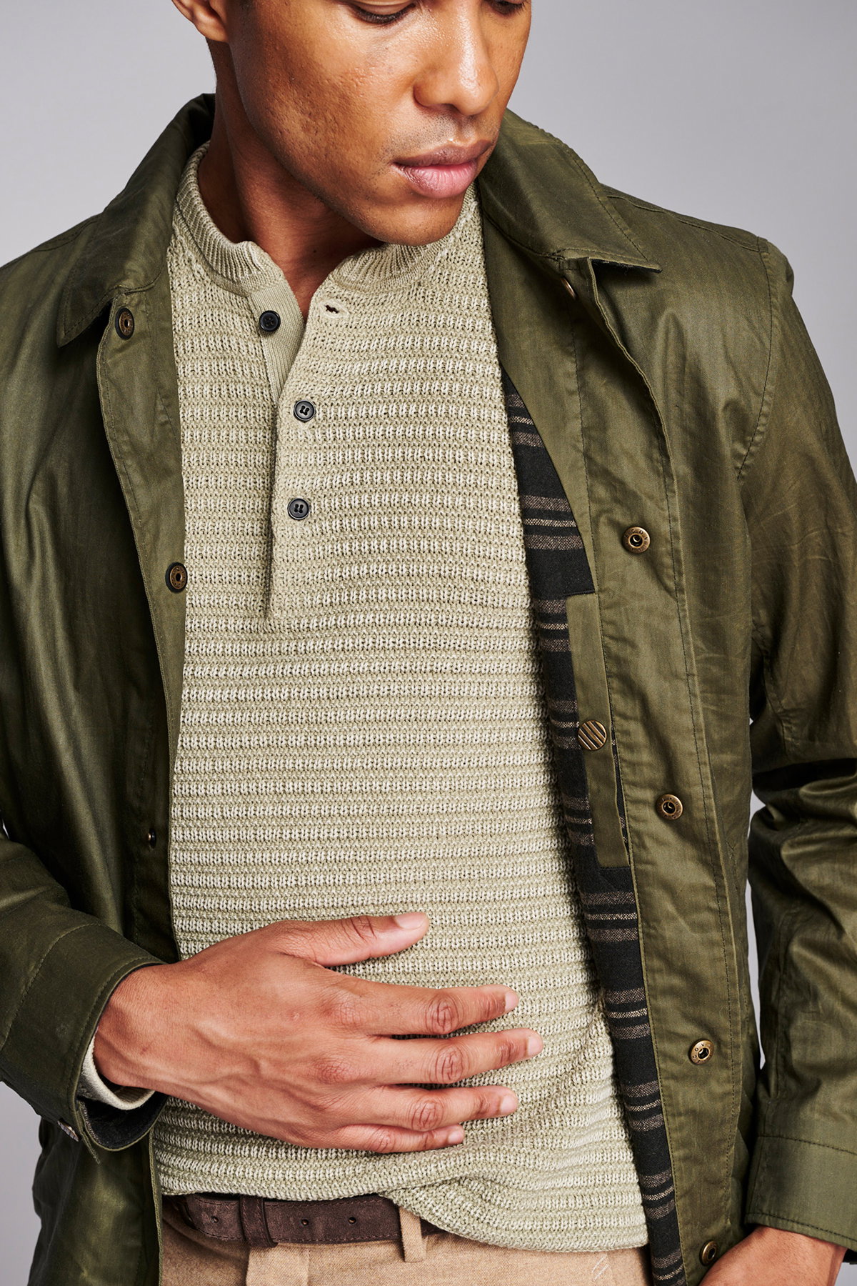 Model wears a dark green waxed jacket over a light green knit henley. 