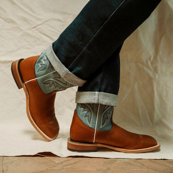 Person wearing jeans and wearing a pair of Tony Lama boots standing in front of a fabric background.