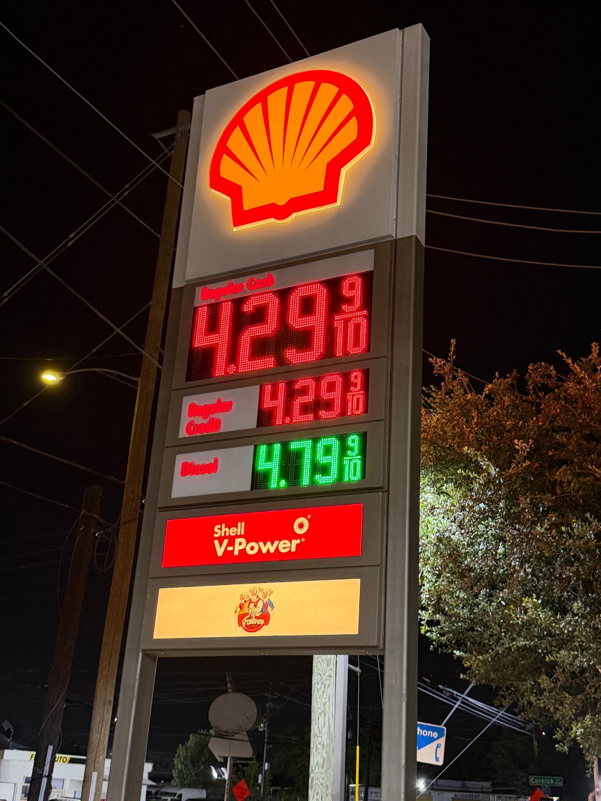 Shell gas station sign with gas prices: "Regular Cash: 4.29 (red numbers); Regular Credit: 4.29 (red numbers); Diesel: 4.79 (green numbers)"; below the prices are signs, one that says "Shell V-Power" and another that has three anthropomorphic cows and the name "Now & Forever"