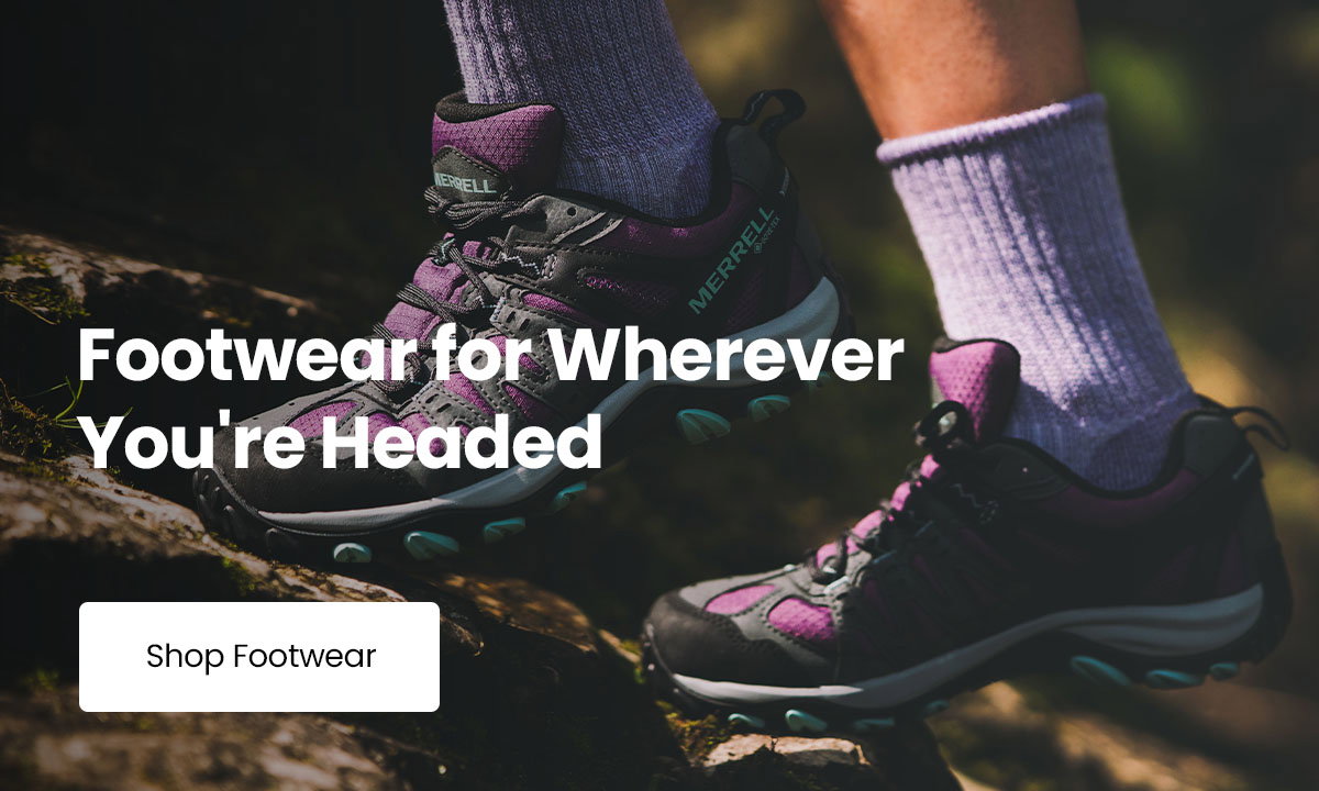 Close-up of purple and black Merrell hiking shoes worn on a rocky forest trail, with the text “Footwear for Wherever You're Headed” and a “Shop Footwear” button displayed over the image.