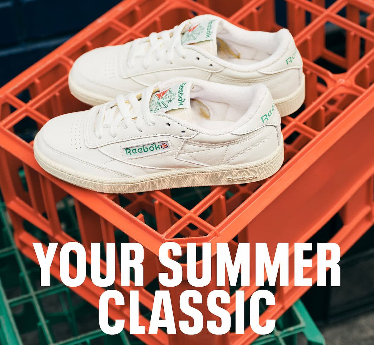 Pair of white Reebok Club C 85 sneakers displayed on stacked orange crates. Text reads: “YOUR SUMMER CLASSIC.”