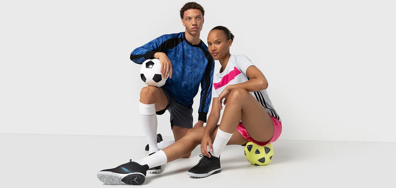 Twee atleten in sportkleding poseren met voetballen. De man draagt een blauw en zwart shirt, zwarte shorts en zwarte schoenen. De vrouw draagt een wit shirt met roze accenten en shorts.