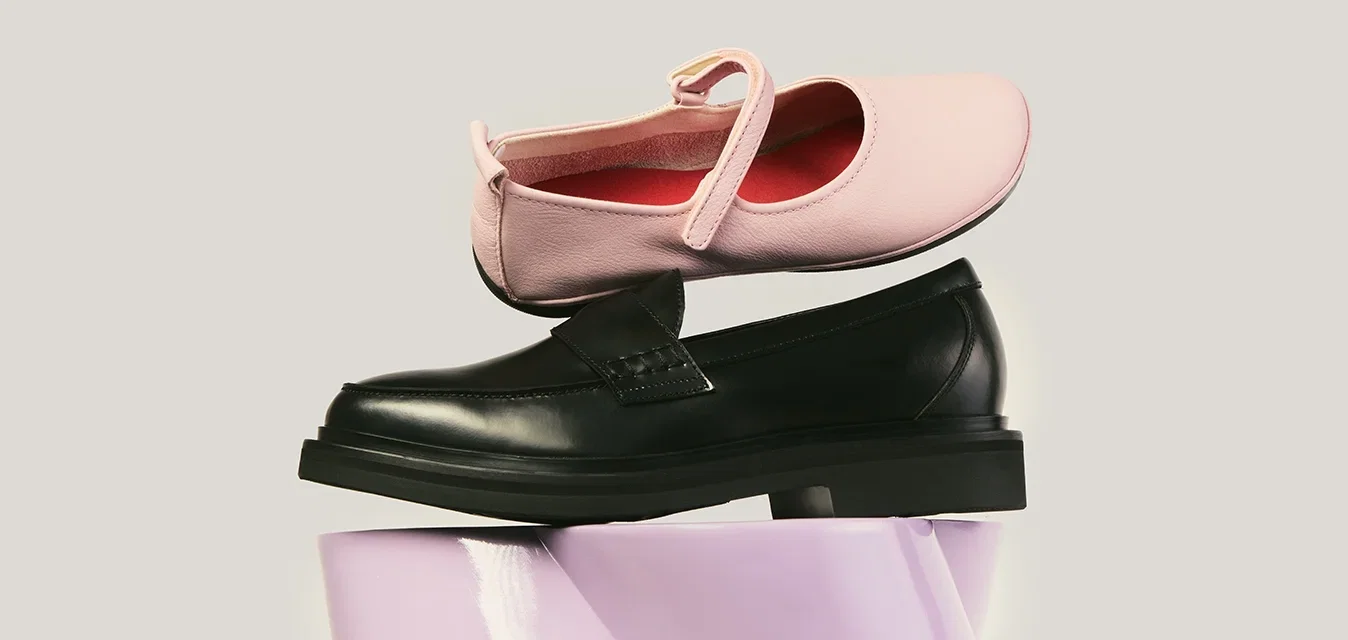 Roze Mary Jane-schoen in evenwicht op een zwarte leren loafer, beide rustend op een glanzend lavendelkleurig oppervlak tegen een effen achtergrond.