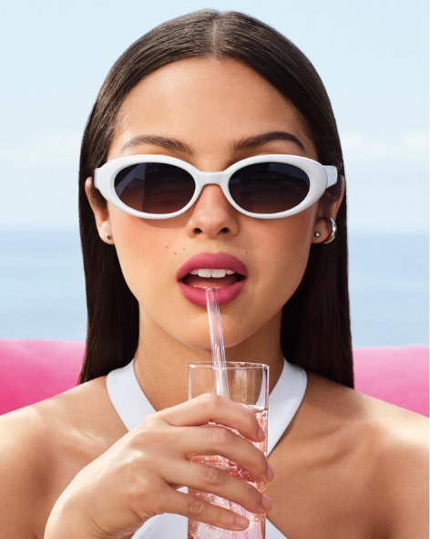 Donna con occhiali da sole bianchi e rossetto rosa mentre beve da un bicchiere con cannuccia