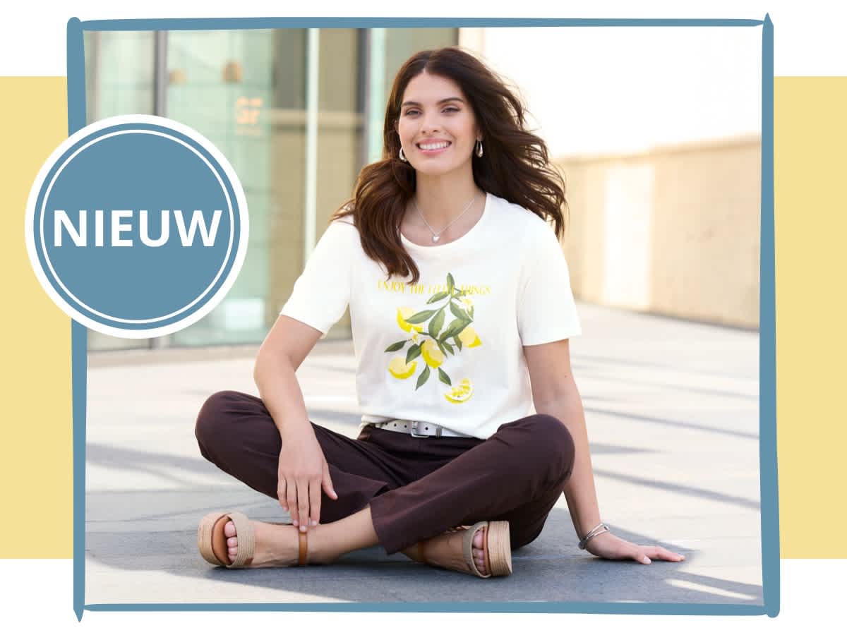 Een model zit buiten op de grond, draagt een wit T-shirt en een bruine broek. Links op de foto is een blauwe cirkel met witte tekst 'NIEUW'.
