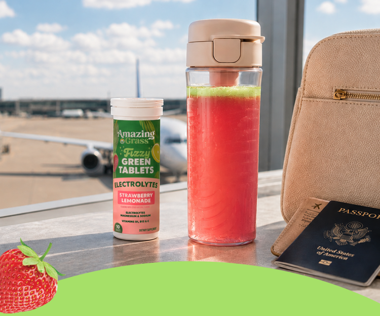 Amazing Grass Fizzy Green Tablets Electrolytes in Strawberry Lemonade flavor displayed beside a pink drink bottle at an airport terminal. A backpack, passport, and boarding pass sit nearby, with an airplane visible outside the window, emphasizing travel-friendly hydration and electrolyte support.