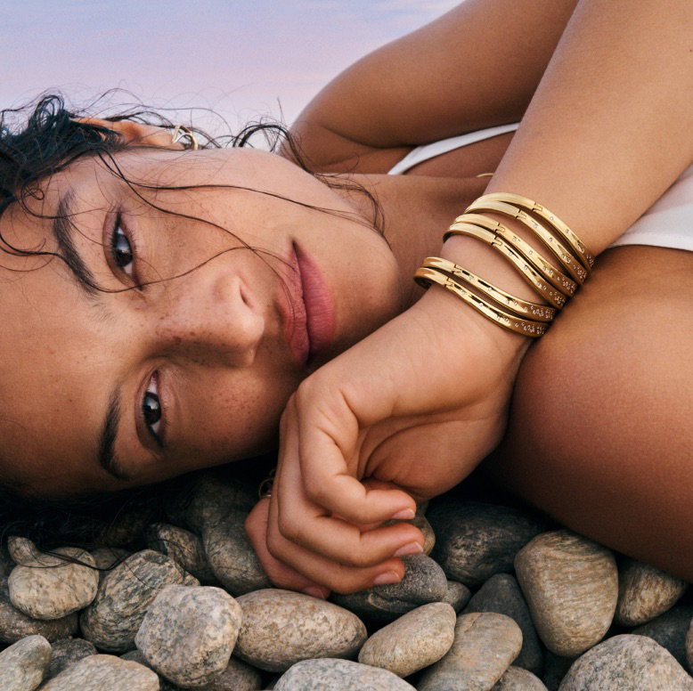 A model rests on pebbles wearing stacked 14k gold-plated bangles.