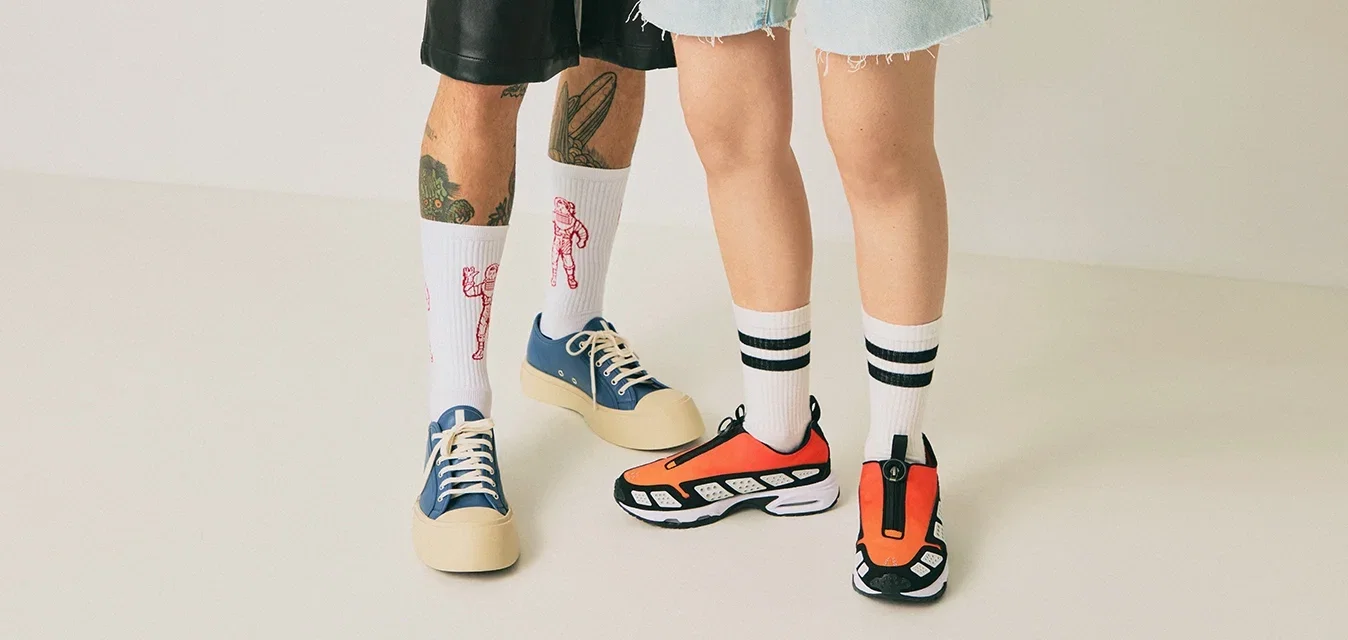 Zwei Personen stehen auf einem hellen Boden, eine trägt schwarze Shorts, blaue Turnschuhe und weiße Socken mit roten Mustern, die andere trägt Jeansshorts, orangefarbene Turnschuhe und gestreifte weiße Socken.