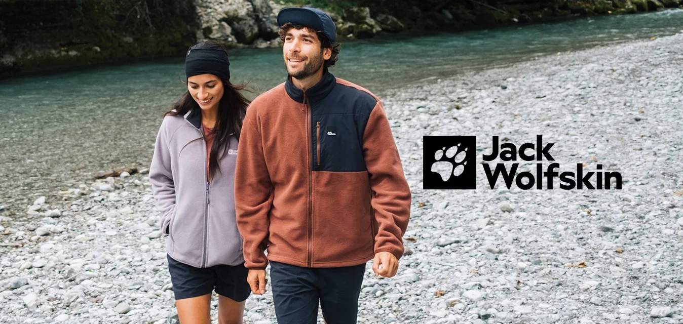 Stel wandelt langs een rotsachtige rivierbedding. Vrouw in een lichtpaarse fleecejas, man in een bruine fleece met zwarte inzet. Beiden in shorts en sneakers.