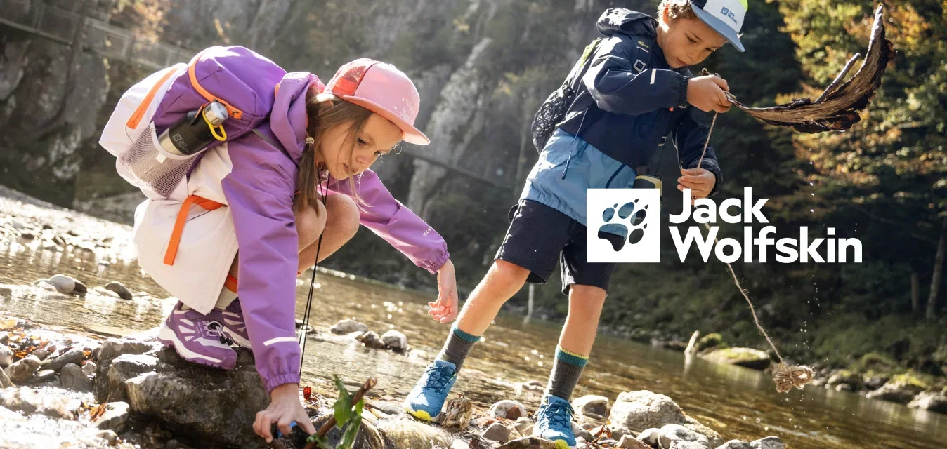 Twee kinderen met rugzakken spelen bij een rotsachtige beek, waarbij ze stokjes en bladeren in het water leggen, met bos en kliffen op de achtergrond.