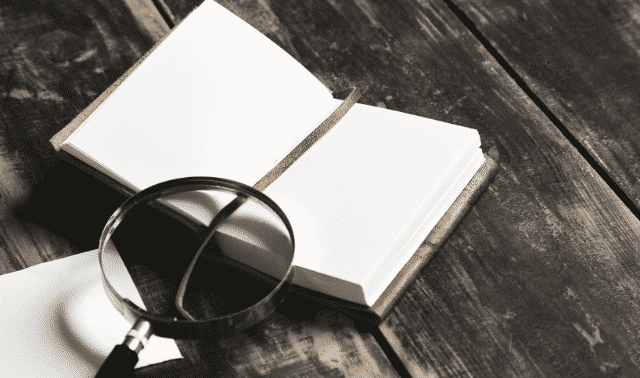 Open blank notebook with a brown cover lies on a weathered wooden surface. A magnifying glass rests on top of the notebook.