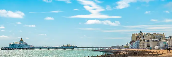 A sunny beach scene with pebbles, a pier, and buildings along the shore under a blue sky.